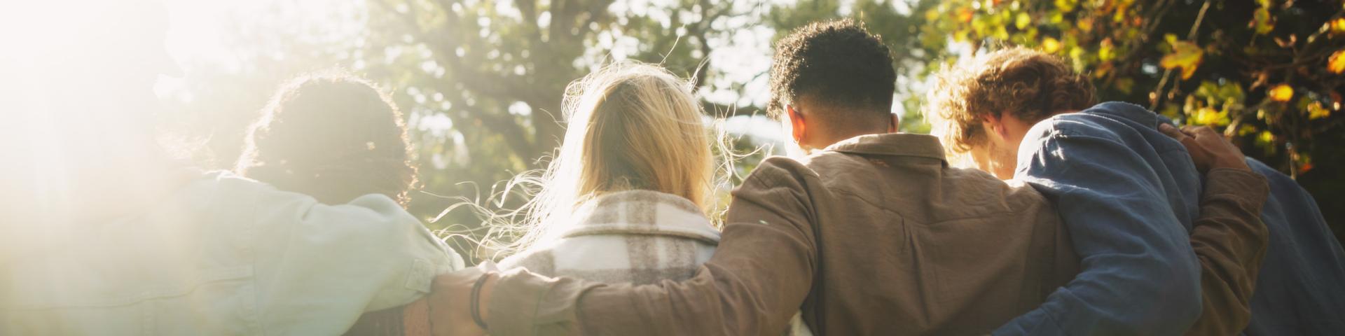 A group of community members hug, standing shoulder to shoulder in a park setting on a nice day, their backs to the camera.