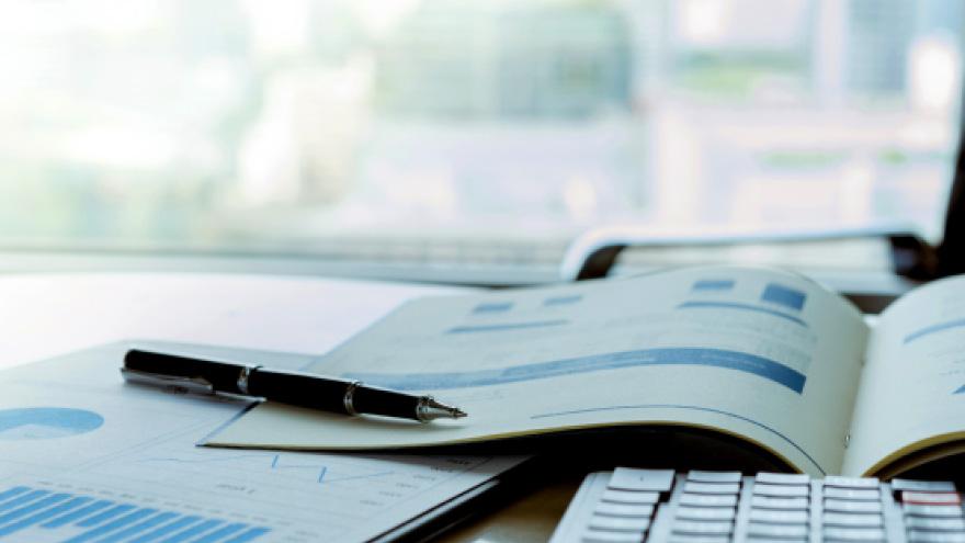 A pen and open report with charts and graphs sprawled across a desk, a blurred window view of the city in the background.